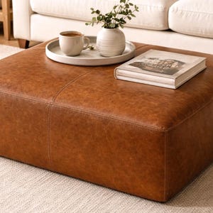 May include: A large, square, brown leather ottoman with visible stitching. A white tray holds a coffee cup, a small white vase with greenery, and a book. The ottoman sits on a light-colored rug, with a white sofa in the background.