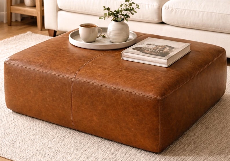 May include: A large, square, brown leather ottoman with visible stitching. A white tray holds a coffee cup, a small white vase with greenery, and a book. The ottoman sits on a light-colored rug, with a white sofa in the background.