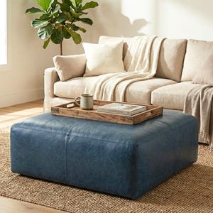 May include: A blue leather ottoman sits on a woven jute rug in front of a beige sofa. A wooden tray with a mug and a book rests on the ottoman. A plant is in the background.