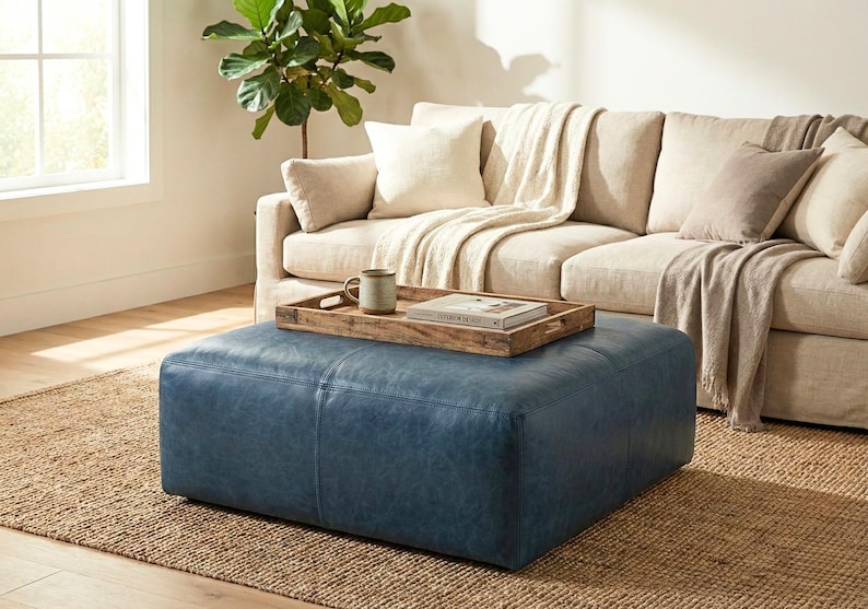 May include: A blue leather ottoman sits on a woven jute rug in front of a beige sofa with pillows and a blanket. A wooden tray with a mug and a book rests on the ottoman. A potted plant is in the background.