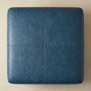 May include: Square, blue ottoman with a textured leather appearance. The top surface is divided into four sections by stitched seams. The ottoman is a solid color and has a simple, modern design.