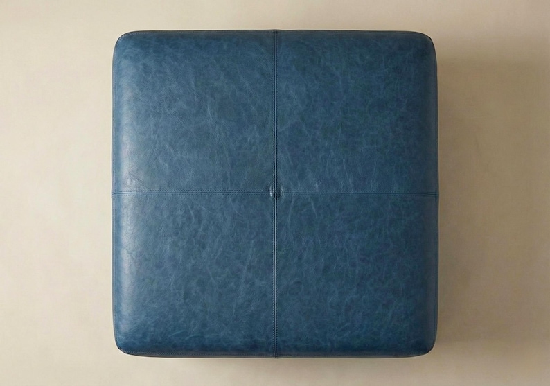 May include: Square, blue ottoman with a textured leather appearance. The top surface is divided into four sections by stitched seams. The ottoman is a solid color and has a simple, modern design.