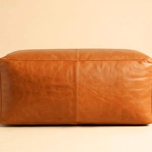 Peut inclure: Un pouf rectangulaire en cuir marron. Le pouf est d'un brun moyen chaud et semble être fait de cuir de haute qualité. Les coutures sont visibles et le design général est simple et moderne.