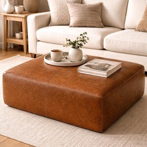 May include: A large, square, brown leather ottoman sits on a light-colored rug in a living room setting. A white sofa with neutral-toned pillows is in the background. A tray with a cup and vase rests on the ottoman, along with a book.