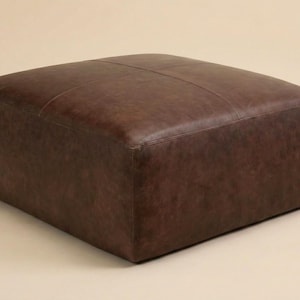May include: A square, brown leather ottoman. The ottoman has a stitched top and a solid, blocky shape. The leather has a slightly distressed appearance, with variations in color and texture. The piece is a neutral, versatile furniture item.