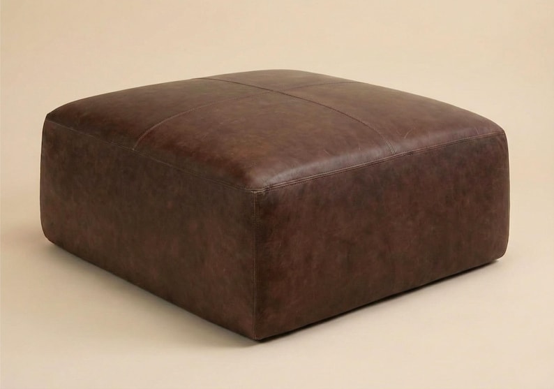 May include: A square, brown leather ottoman. The ottoman has a stitched top and a solid, blocky shape. The leather has a slightly distressed appearance, with variations in color and texture. The piece is a neutral, versatile furniture item.