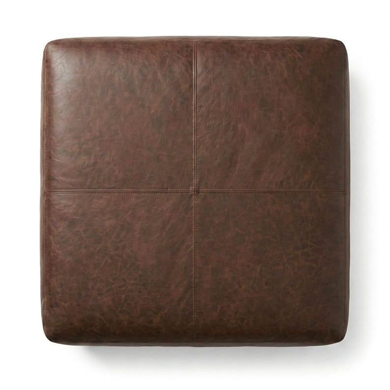 May include: A square, brown leather ottoman with a textured surface. The ottoman is divided into four sections by stitched seams. The color is a rich, dark brown.