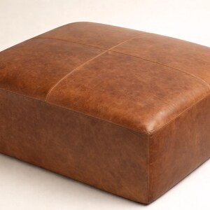 May include: A square, brown leather ottoman. The top is divided into four sections by stitched lines. The leather has a slightly distressed appearance. The ottoman is a low, rectangular shape.