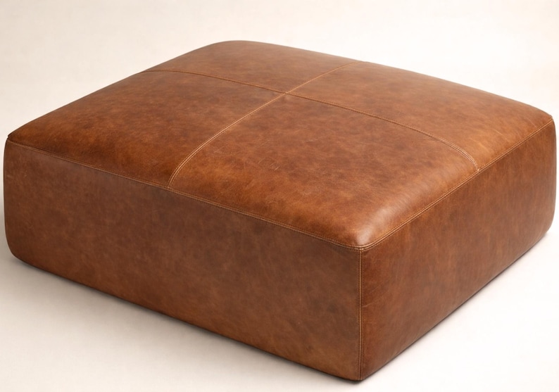 May include: A square, brown leather ottoman. The top is divided into four sections by stitched lines. The leather has a slightly distressed appearance. The ottoman is a low, rectangular shape.