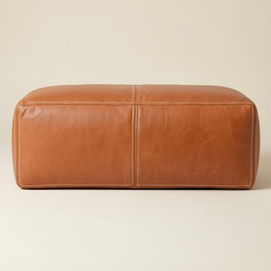 Peut inclure: Un pouf rectangulaire en cuir marron clair avec des coutures visibles. Le pouf a un aspect rembourré et une texture lisse. Les coutures sont d'une teinte plus claire que le cuir, créant un contraste subtil.