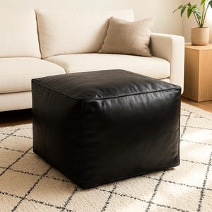 Könnte beinhalten: Ein quadratischer, schwarzer Lederhocker. Das würfelförmige Möbelstück steht auf einem cremefarbenen Teppich mit geometrischem Muster. Ein beiges Sofa und ein Beistelltisch mit einer Pflanze befinden sich im Hintergrund.