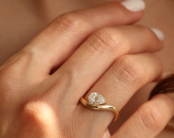 May include: A gold ring with a pear-shaped diamond. The ring's band curves gracefully, cradling the diamond. The ring is shown on a hand with white nail polish.