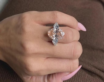 May include: A large, ornate ring featuring a central oval-cut, peach-colored gemstone, flanked by two smaller, pear-shaped, clear gemstones. The ring is set in a silver or white metal, creating a striking contrast with the skin tone.