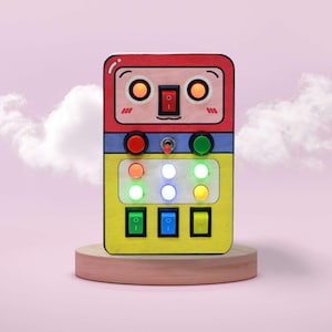 May include: A brightly coloured wooden toy with a robot face design. It has red, blue, and yellow sections with buttons, switches, and lights. The toy is on a small wooden stand against a pink background with a cloud.