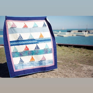 May include: A handmade quilt featuring a nautical theme. The quilt displays a pattern of sailboats in various colors, including red, blue, and orange, set against a white and blue background. The quilt is bordered with a dark blue fabric.