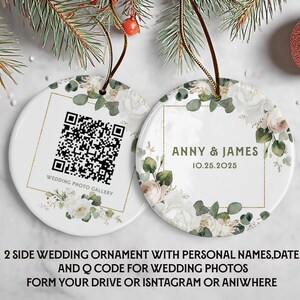 May include: Two white ceramic wedding ornaments with floral designs and gold trim. One has a QR code and the text "WEDDING PHOTO GALLERY". The other says "ANNY & JAMES 10.25.2025". Both have a brown string for hanging. The ornaments are surrounded by greenery.