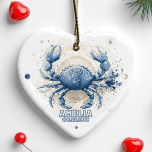May include: A heart-shaped ceramic ornament featuring a blue crab design, the word "AMELIA" and the date 06.23.1987. The ornament hangs from a gold cord. The background includes pine cones, red heart-shaped decorations, and greenery.