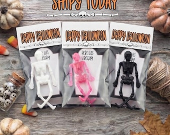 May include: Three clear plastic bags containing white, pink, and black skeleton figures. Each bag has a header card with the text "Happy Halloween" and a tombstone graphic with the text "Here lies stretchy". The skeletons are in a seated position.