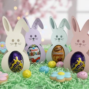 May include: Easter-themed display featuring four pastel-colored bunny-shaped holders, each cradling a chocolate egg. The scene includes decorative cookies, colorful candies, and artificial grass, creating a festive holiday arrangement.