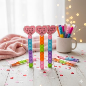 May include: Three colorful pop-it toys with heart-shaped toppers that read "You Make My Heart Pop." The toys are in shades of blue, orange, green, and purple. Other pop-it toys and pens are in the background.