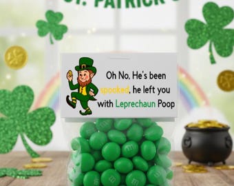May include: A clear cellophane bag filled with green candies, labeled "Leprechaun Poop," sits on a wooden surface. The image includes a St. Patrick's Day banner, shamrocks, gold coins, and a pot of gold.