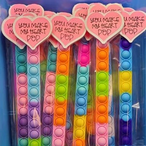 May include: A collection of colorful, rainbow-colored pop-it fidget toys. Each toy is packaged in a clear bag and topped with a pink heart-shaped tag that reads "You Make My Heart Pop."