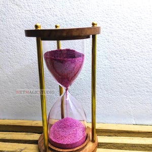 May include: An hourglass with a wooden frame and brass-colored supports. The top and bottom are made of wood, and the hourglass contains pink sand. The text "METMAGICSTUDIO" is visible on the side.