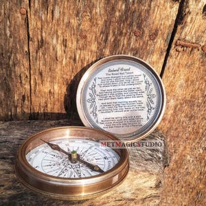 May include: A round brass compass with a white dial featuring black cardinal directions and a needle. The lid is engraved with the poem The Road Not Taken by Robert Frost, framed by leaf patterns. The text METMAGICSTUDIO is visible on the side.