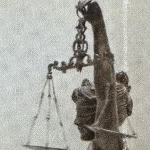 Puede incluir: Una escultura de tonos oscuros de una figura que sostiene una balanza en alto. La balanza presenta una barra transversal ornamentada suspendida de un brazo vertical, con cadenas finas que conectan a un plato de pesaje triangular.