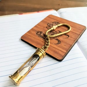 May include: A brass hourglass keychain featuring a cylindrical metal frame with clear glass and white sand inside. The hourglass is attached to a brass chain and a carabiner-style clasp, resting on a square wooden piece with a dark engraved design.
