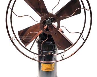 May include: A vintage-style desk fan with four dark copper-toned blades enclosed in a circular wire cage. The base is a cylindrical black metal stand featuring a small rectangular brass plaque with the text East India Company engraved on it.