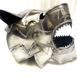 Könnte beinhalten: Ein polierter, silberfarbener Metallhelm in Form eines Wolfskopfes mit offenem Kiefer und scharfen, gezackten Zähnen. Der Helm besteht aus überlappenden, beweglichen Metallplatten, die ein geschichtetes und segmentiertes Erscheinungsbild erzeugen.