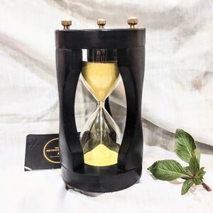 May include: An hourglass with a black frame and yellow sand. The frame features three small brass-colored knobs on the top. A black rectangular card with the text METMAGI is positioned to the left, and a small green leafy sprig sits to the right.