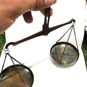 Puede incluir: Una balanza de mano con acabado metálico oscuro y dos platos circulares suspendidos por cadenas finas de una barra horizontal central. La estructura está hecha de metal oscuro y desgastado, con un diseño clásico de pesaje.