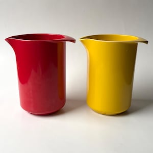 May include: Two cylindrical vases, one red and one yellow, with a curved spout. The vases have a glossy finish and are set against a white background. The vases are similar in shape and size.