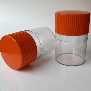 May include: Two clear plastic containers with bright orange lids. One container is upright, the other is on its side. The lids have a ridged texture. The containers are on a white surface.