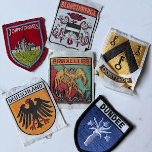 May include: A collection of six embroidered fabric patches featuring various heraldic designs and city names including John O'Groats, Blankenberge, Bruxelles, Oostende, Deutschland, and Dundee, each with distinct colours and detailed stitching.
