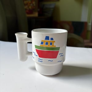 May include: White ceramic mug featuring a boat design. The boat is red with yellow and blue details, and green trim, sailing on blue waves. The mug has a unique handle design.