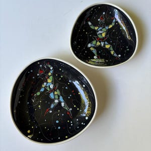 May include: Two irregular, rounded ceramic bowls with a glossy black interior featuring a colourful, mosaic-style harlequin figure in the centre. The figures are depicted in dynamic poses surrounded by thin white swirling lines and small dots.