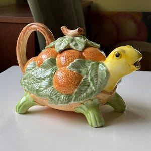 May include: A ceramic teapot in the shape of a turtle. The turtle has a yellow head and green legs. The shell is decorated with orange fruit and green leaves. The handle and spout are light brown.