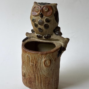 May include: A ceramic planter shaped like a tree stump with a textured brown finish and a sculpted owl perched on top. The owl features large circular eyes, dark brown spots on its chest, and is positioned above a hollow opening in the stump.