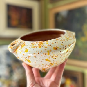 May include: A cream-coloured ceramic bowl with a speckled pattern of yellow and orange spots. The interior is brown. The bowl has two handles and three small feet.