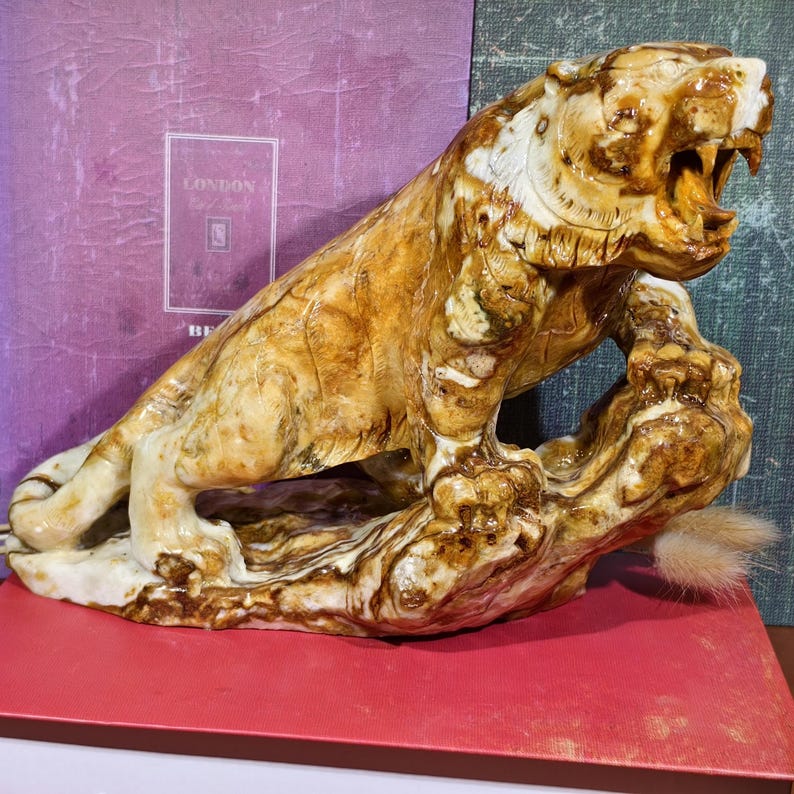 May include: A detailed sculpture of a tiger in a roaring pose, crafted from a light brown material with darker brown veining. The tiger is perched on a textured, brown base, with its mouth open, revealing teeth. The sculpture has a glossy finish.