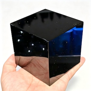 May include: A solid, smooth, black cube with a highly reflective, polished surface. The object features sharp, clean edges and corners, creating a geometric shape that mirrors its surroundings on its dark, glossy faces in a bright environment.