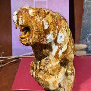 May include: A detailed sculpture of a roaring tiger, carved from a stone with brown, tan, and white hues. The tiger is depicted in a dynamic pose, with its mouth open, showcasing its teeth. The sculpture is set on a red surface.