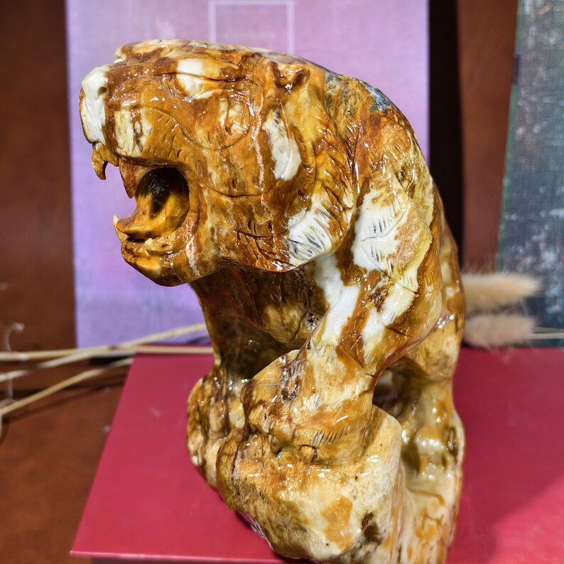 May include: A detailed sculpture of a roaring tiger, carved from a stone with brown, tan, and white hues. The tiger is depicted in a dynamic pose, with its mouth open, showcasing its teeth. The sculpture is set on a red surface.