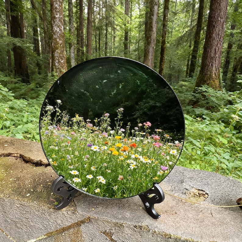May include: A round, dark green mirror reflecting a vibrant wildflower meadow. The mirror is displayed on a black metal stand, set against a backdrop of a lush forest. The wildflowers include white, yellow, orange, and pink blooms.