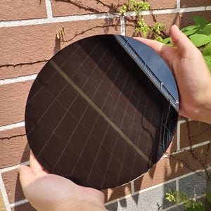 Könnte beinhalten: Ein großes, rundes, schwarzes Objekt mit einem Rastermuster wird vor einer Backsteinmauer gehalten. Das Objekt reflektiert ein Gebäude und den Himmel. Das Objekt wird von zwei Händen gehalten.