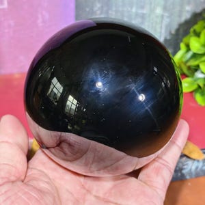 Op de afbeelding: Een gepolijste zwarte obsidiaan bol, die licht en omringende objecten reflecteert. De bol wordt in een hand gehouden en toont zijn gladde, glanzende oppervlak. Het is een decoratief item.