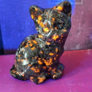 Könnte beinhalten: Kleine, gefleckte Katzenfigur in sitzender Position. Die Skulptur ist überwiegend dunkelgrau mit leuchtend orangefarbenen und schwarzen Flecken. Augen und Ohren der Katze sind deutlich definiert, und das Gesamtdesign ist glatt und poliert.